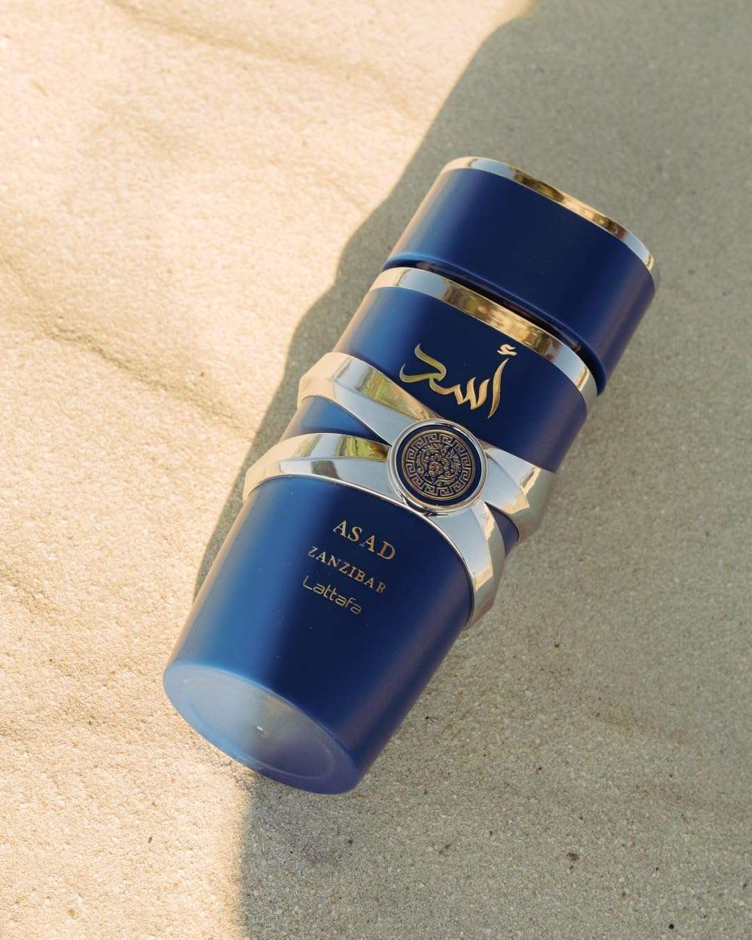 Lattafa Asad Zanzibar perfume men in a sleek blue and gold bottle placed on sandy surface Lattafa Asad Zanzibar perfume men in a sleek blue and gold bottle placed on sandy surface