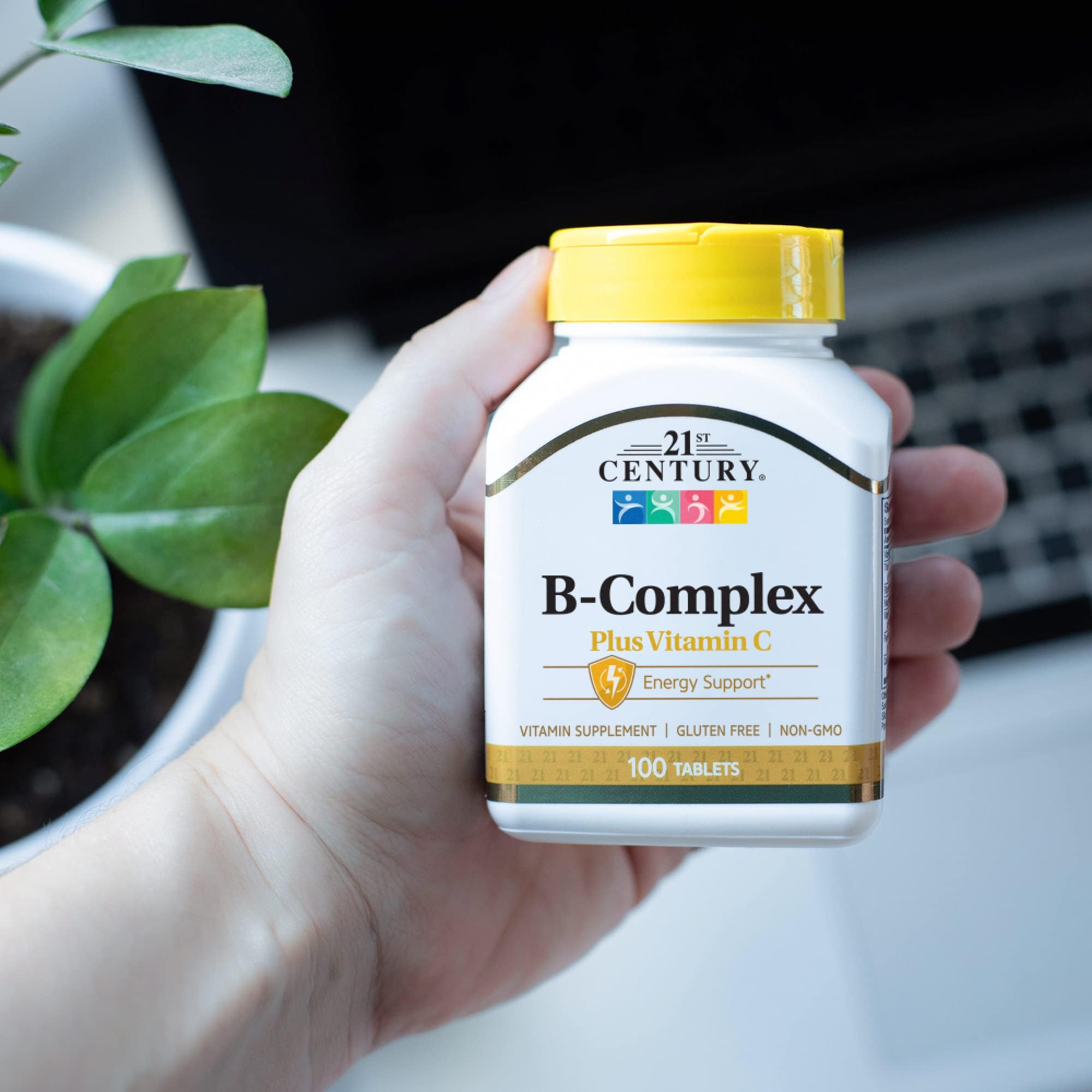 21st Century B-Complex bottle with yellow cap held in hand near plant and laptop, B-Complex with Vitamin C supplements UAE