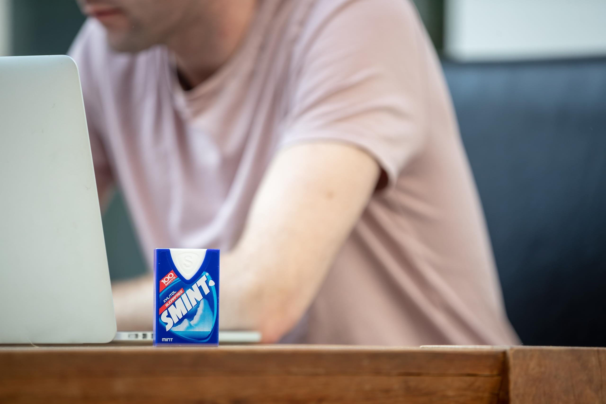 Smint sugarfree mints pack on a wooden desk next to a laptop with a person working blurred in background