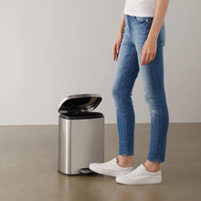 Brushed stainless steel trash can with foot pedal, lid open as someone steps