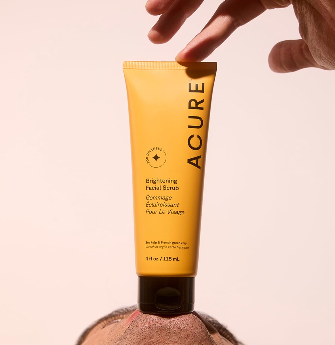 Acure brightening facial scrub in yellow tube held above a person's face for skincare routine
