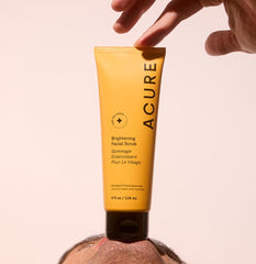 Acure brightening facial scrub in yellow tube held above a person's face for skincare routine