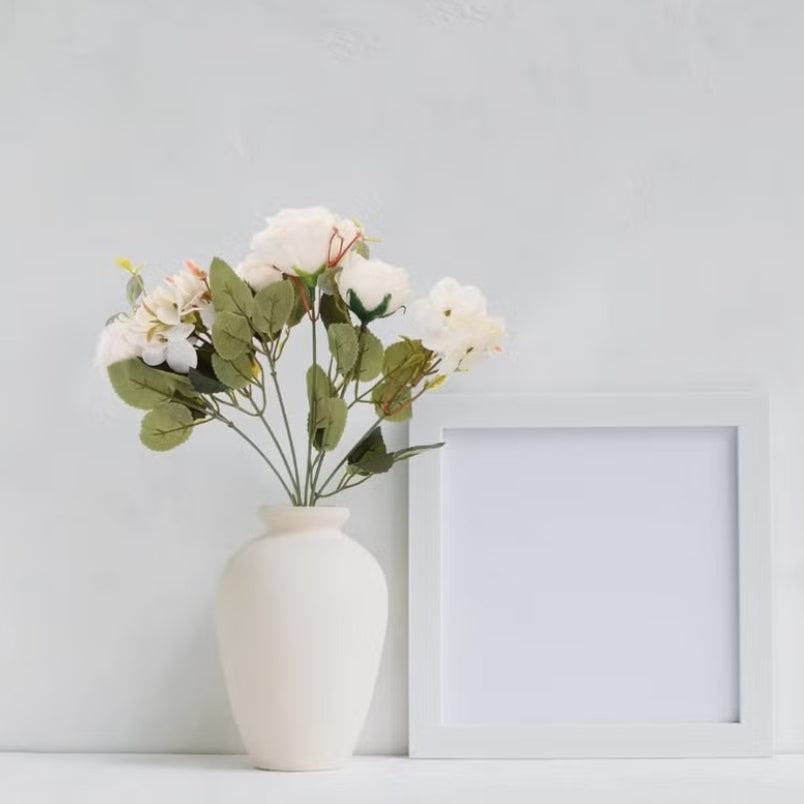 Huitich artificial mini white roses arranged in a white ceramic vase next to a blank white photo frame
