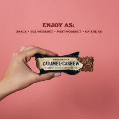 Hand holding partially unwrapped Barebells protein bars caramel cashew with crunchy nut coating on pink background