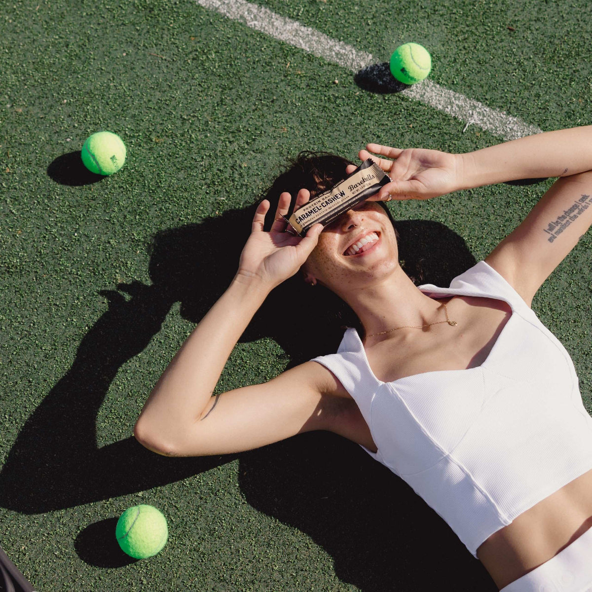 Smiling woman lying on tennis court holding Barebells protein bars caramel cashew bar over eyes with tennis balls nearby