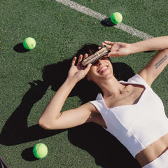 Smiling woman lying on tennis court holding Barebells protein bars caramel cashew bar over eyes with tennis balls nearby