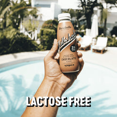 Hand holding Barebells Chocolate 24g protein milkshake by a pool labeled lactose free Barebells protein milkshake UAE