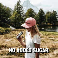 Woman in pink Barebells cap holding a high protein cookies and cream bar by a mountain lake