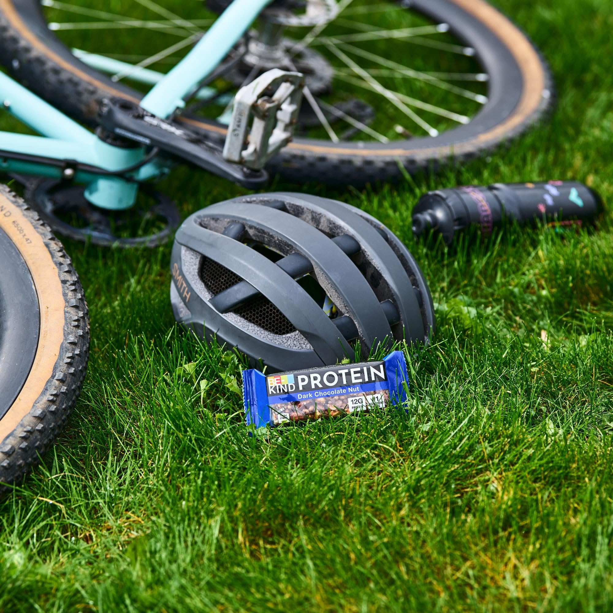 BE KIND dark chocolate protein bars beside bike helmet and water bottle on grass for outdoor rides