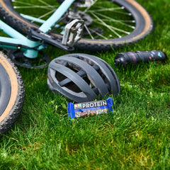 BE KIND dark chocolate protein bars beside bike helmet and water bottle on grass for outdoor rides