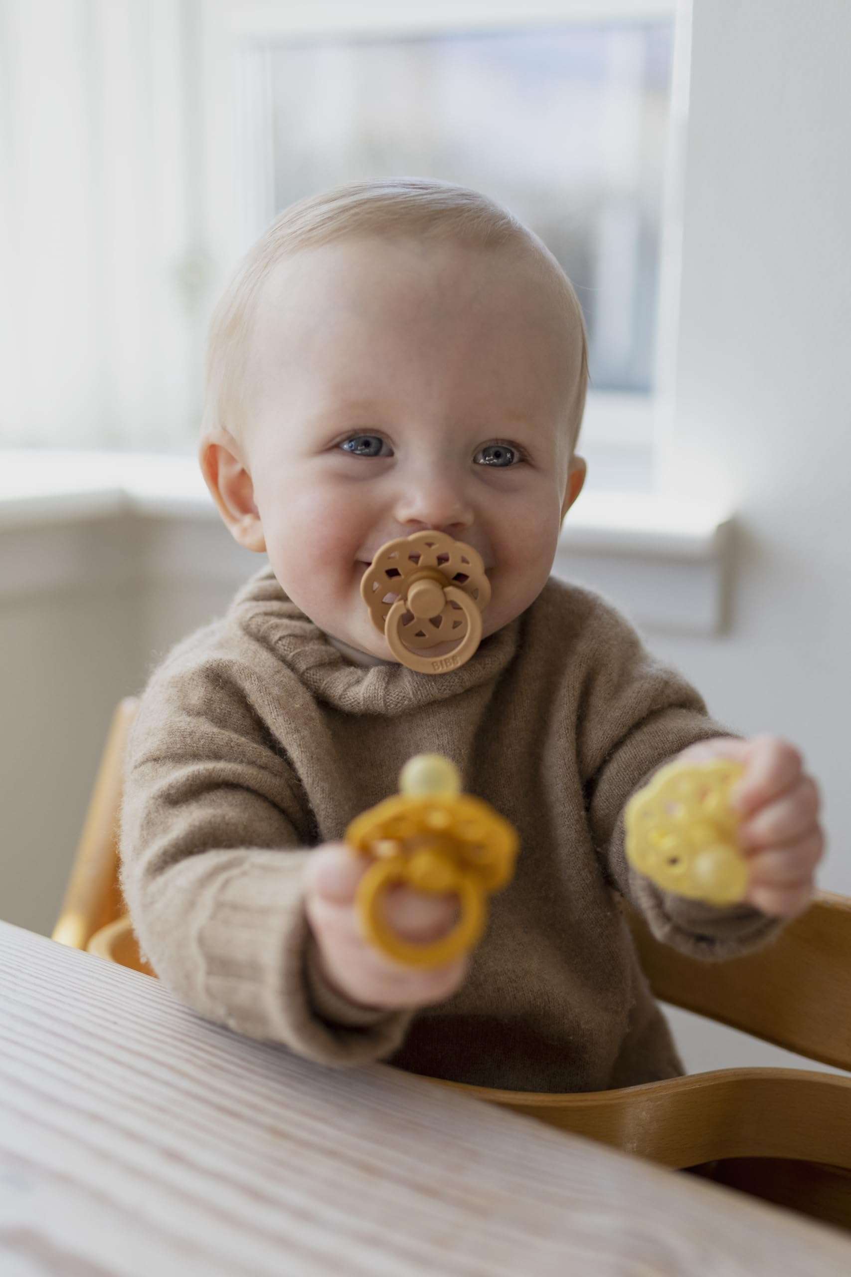 Baby with Bibs Boheme natural rubber pacifier UAE in mouth holding two yellow pacifiers while seated at table