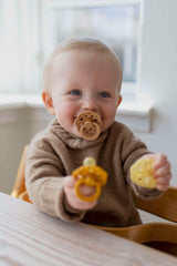 Baby with Bibs Boheme natural rubber pacifier UAE in mouth holding two yellow pacifiers while seated at table