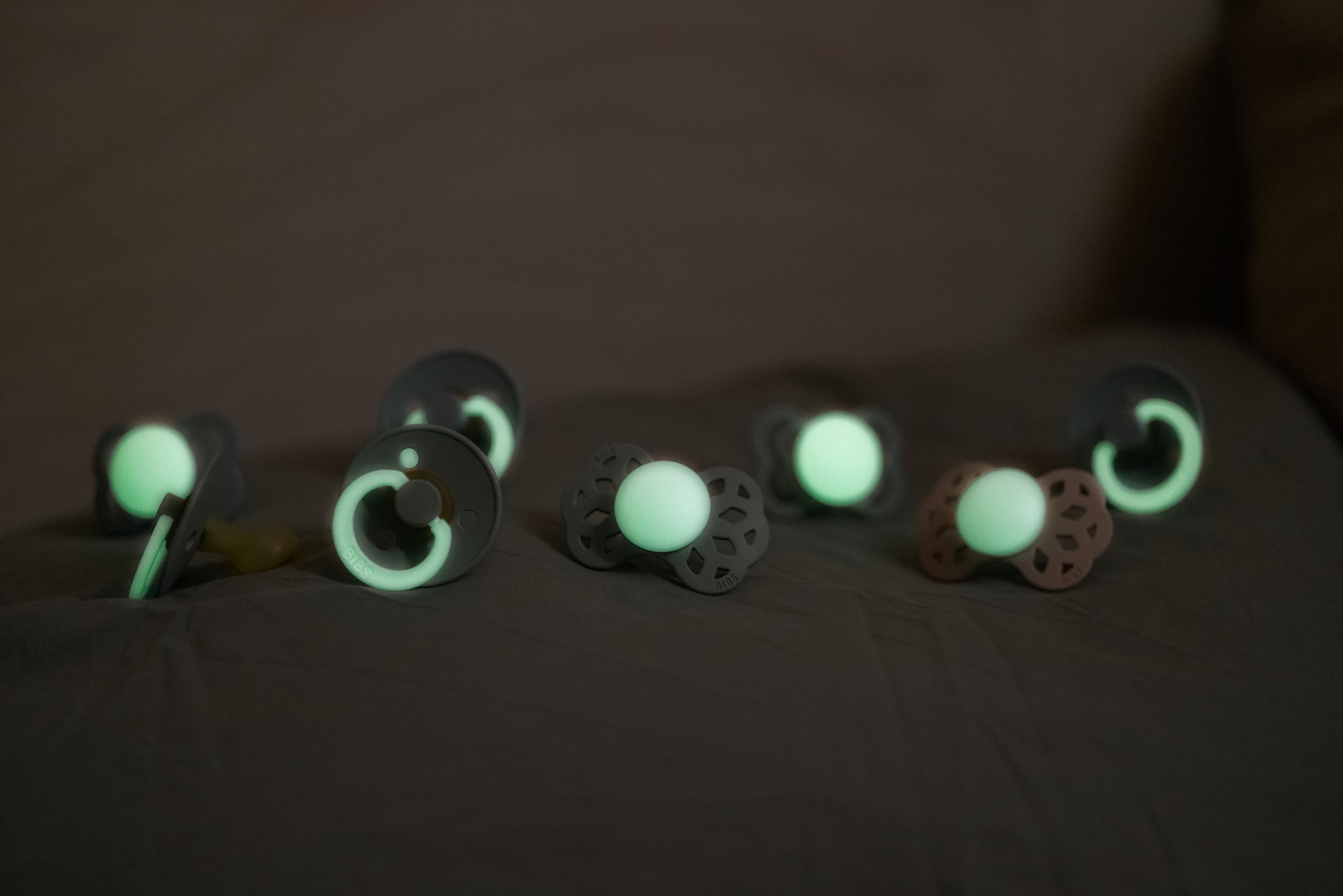 Assorted glowing pacifiers on dark fabric showcasing glow in the dark baby pacifier designs from Visit the Bibs Store