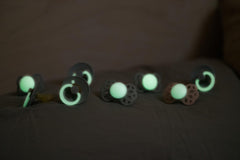 Assorted glowing pacifiers on dark fabric showcasing glow in the dark baby pacifier designs from Visit the Bibs Store