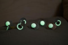 Pack of soft silicone glow in the dark baby pacifier set glowing green on dark nursery bedding for easy finding