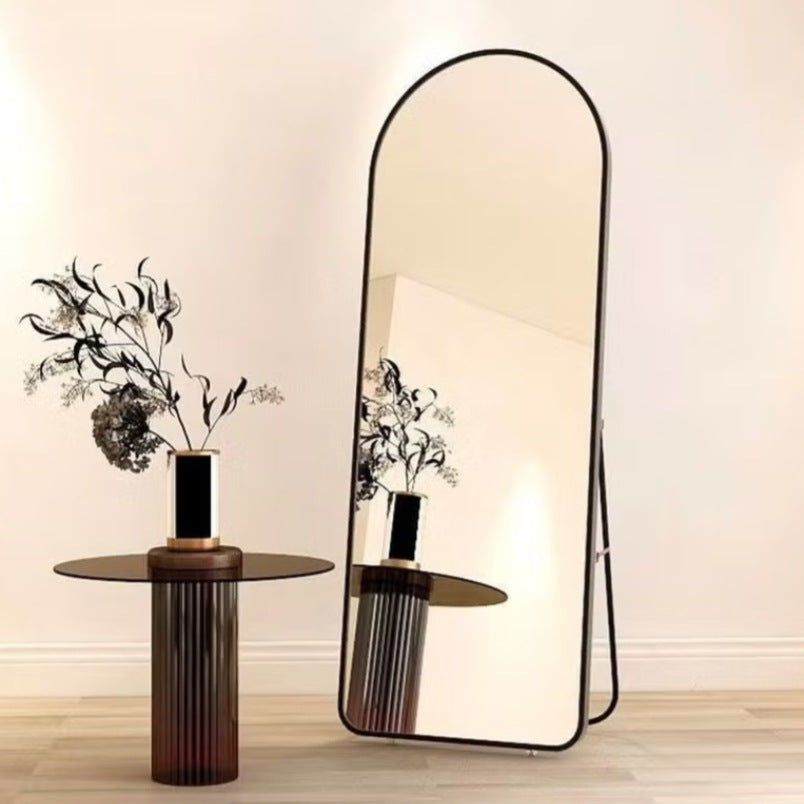 Full-length black aluminum frame mirror with rounded top standing next to modern side table with vase