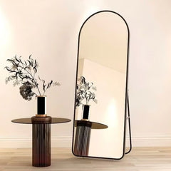 Full-length black aluminum frame mirror with rounded top standing next to modern side table with vase