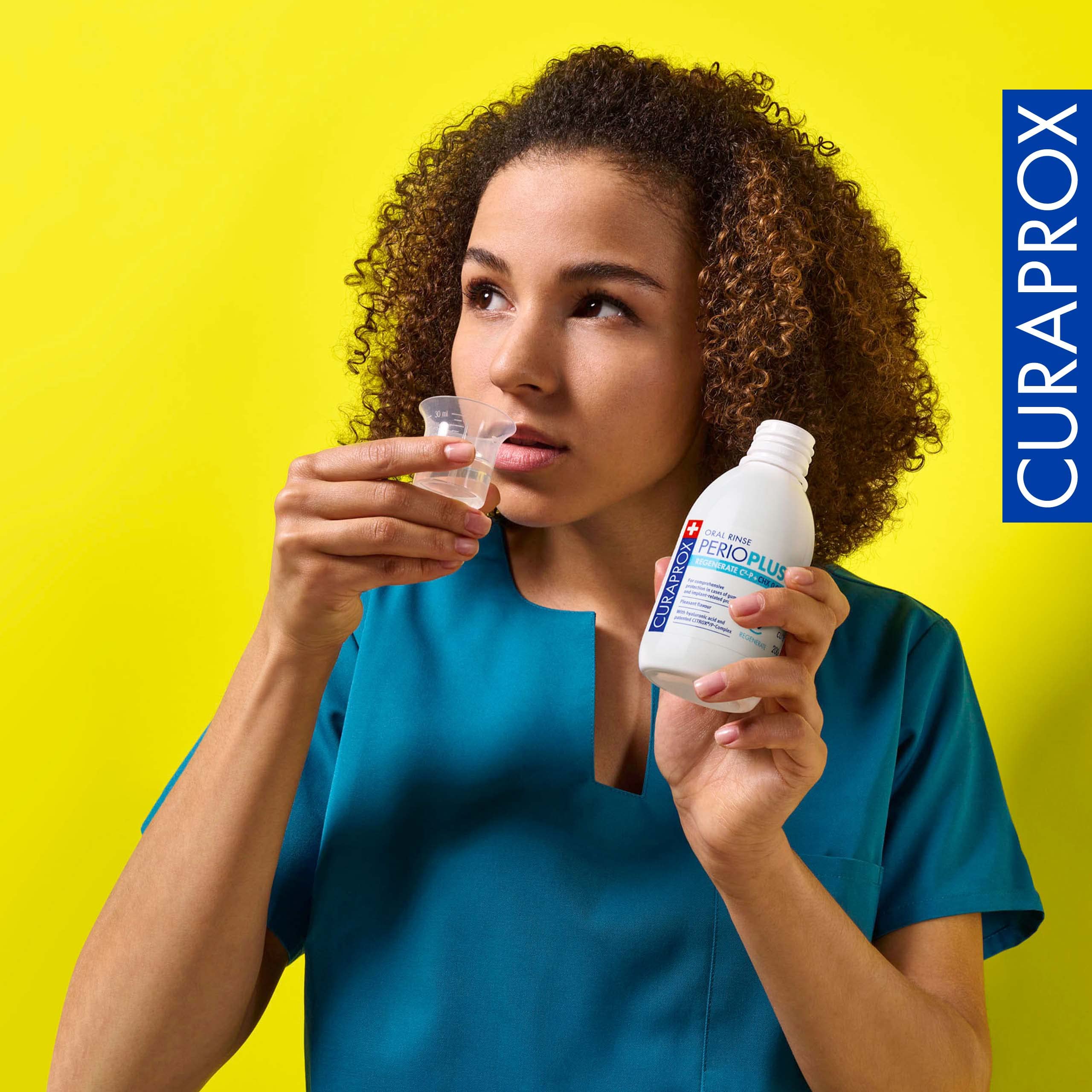 Dental professional in teal scrubs holding Curaprox Perio Plus Regenerate mouthwash bottle and dosing cup against yellow background