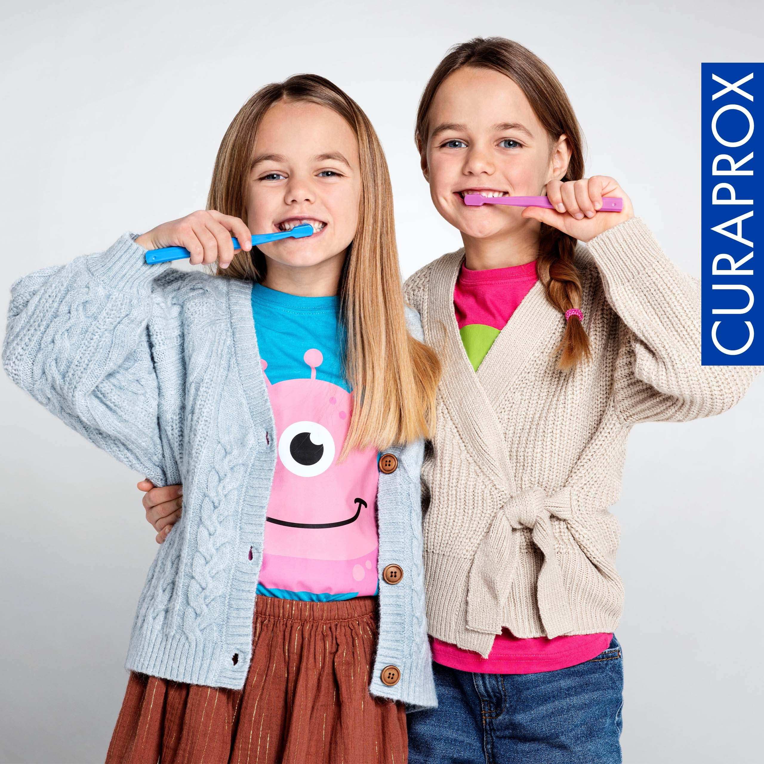 Two smiling girls brushing teeth with colorful toothbrushes promoting kids fluoride toothpaste strawberry flavor by Curaprox