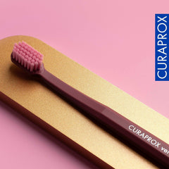 Curaprox maroon and pink ultra soft toothbrush for sensitive gums resting on gold and pink background