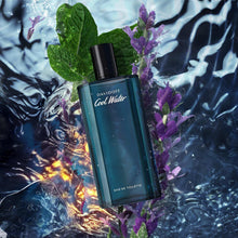 Teal eau de toilette bottle with black cap among green leaves and purple flowers