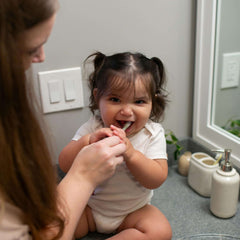 Dr Browns infant toothbrush 0-3 years being used by parent to brush smiling toddler seated on bathroom counter