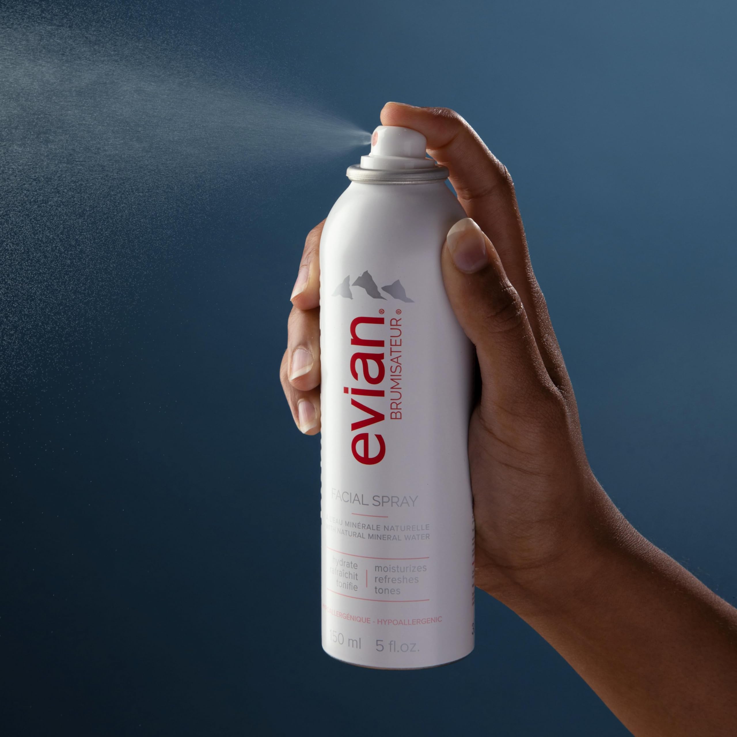 Hand spraying Evian facial spray 5 oz with fine mist against blue background