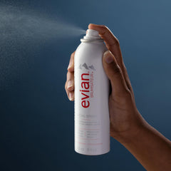 Hand spraying Evian facial spray 5 oz with fine mist against blue background