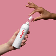 Evian facial spray 5 oz held by two hands with pink cap removed on a pink background
