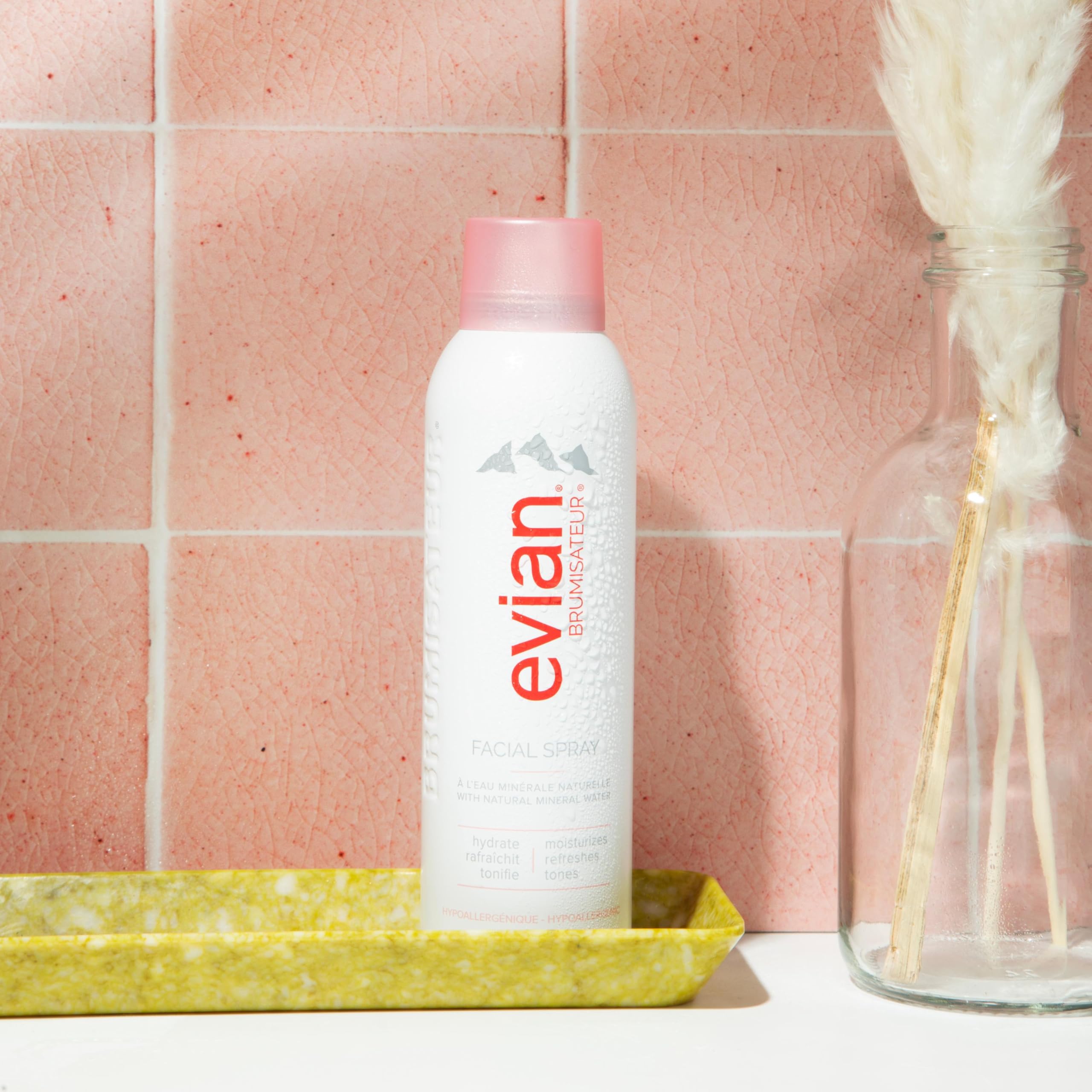 Evian facial spray 5 oz bottle with pink cap standing on yellow tray against pink tiled wall
