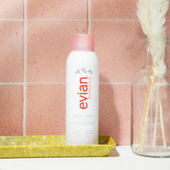 Evian facial spray 5 oz bottle with pink cap standing on yellow tray against pink tiled wall