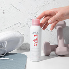 Evian facial spray 5 oz bottle next to white sneaker, yoga mat, and pink dumbbells with hand reaching