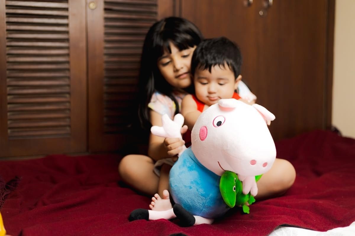 George Buddy soft toy 19x10cm held by a toddler with an older sibling in the background on a red blanket