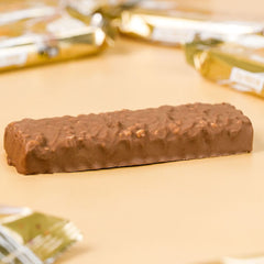 Grenade Carb Killa protein bar unwrapped chocolate coated with nut pieces on beige surface with gold wrappers