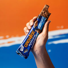 Hand holding bitten Grenade Oreo protein bars chocolate coated bar in blue wrapper on orange background