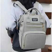 Light gray maternity travel backpack carried by person, with front zip pocket and gold zippers