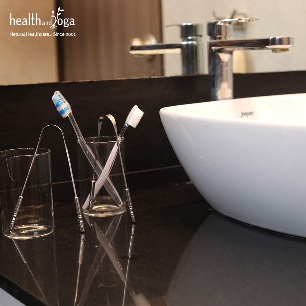 HealthAndYoga metal tongue cleaner on a bathroom countertop beside toothbrushes and a white sink stainless steel oral scraper