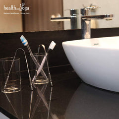 HealthAndYoga metal tongue cleaner on a bathroom countertop beside toothbrushes and a white sink stainless steel oral scraper