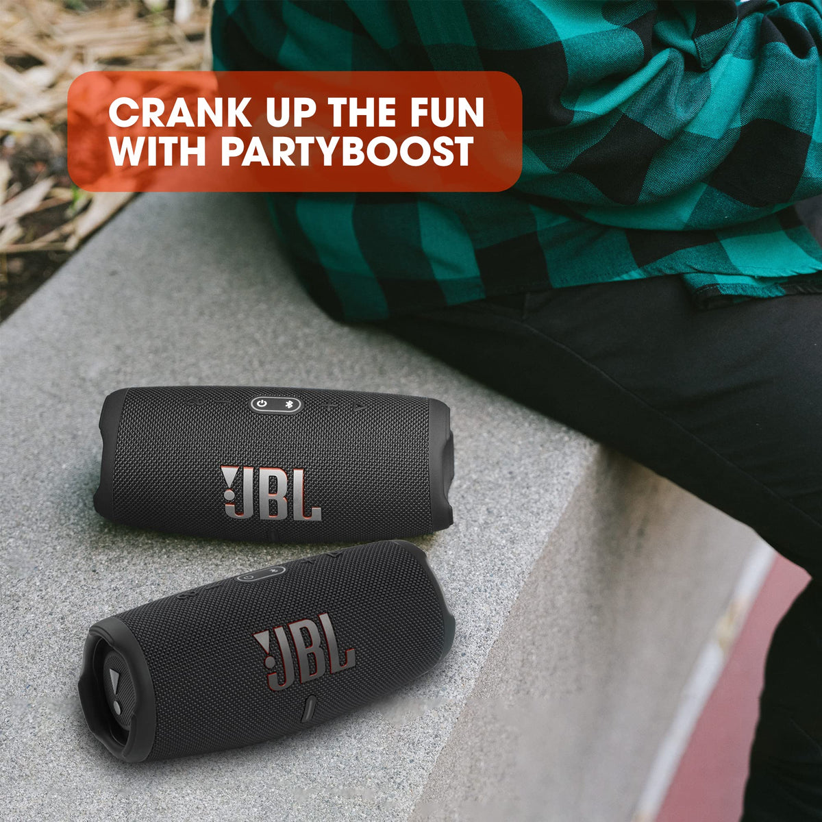Two black JBL Charge 5 portable Bluetooth speakers placed on a concrete surface with a person wearing a green and black checkered shirt nearby