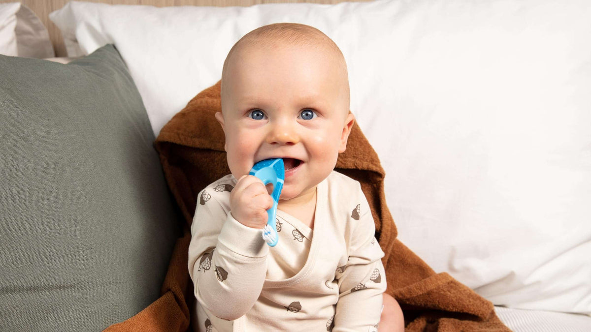 Jordan soft bristle toothbrush for infants blue teether held by smiling baby sitting on bed demonstrating gentle gum cleaning