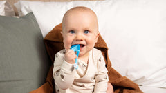 Jordan soft bristle toothbrush for infants blue teether held by smiling baby sitting on bed demonstrating gentle gum cleaning