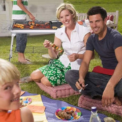Family enjoying a picnic with a large outdoor silver BBQ grill cooking skewers and meat in the background