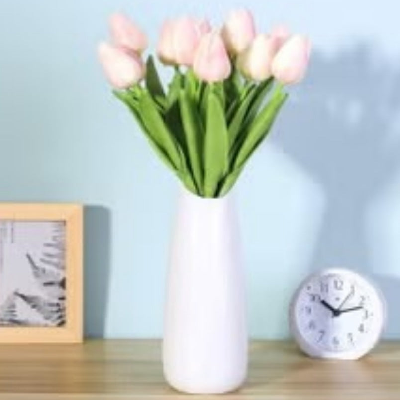 Light pink silk tulips bouquet arranged in a white vase on a wooden table with clock and frame