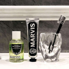 Marvis strong mint mouthwash bottle next to Marvis black licorice toothpaste tube and toothbrush in glass on bathroom counter