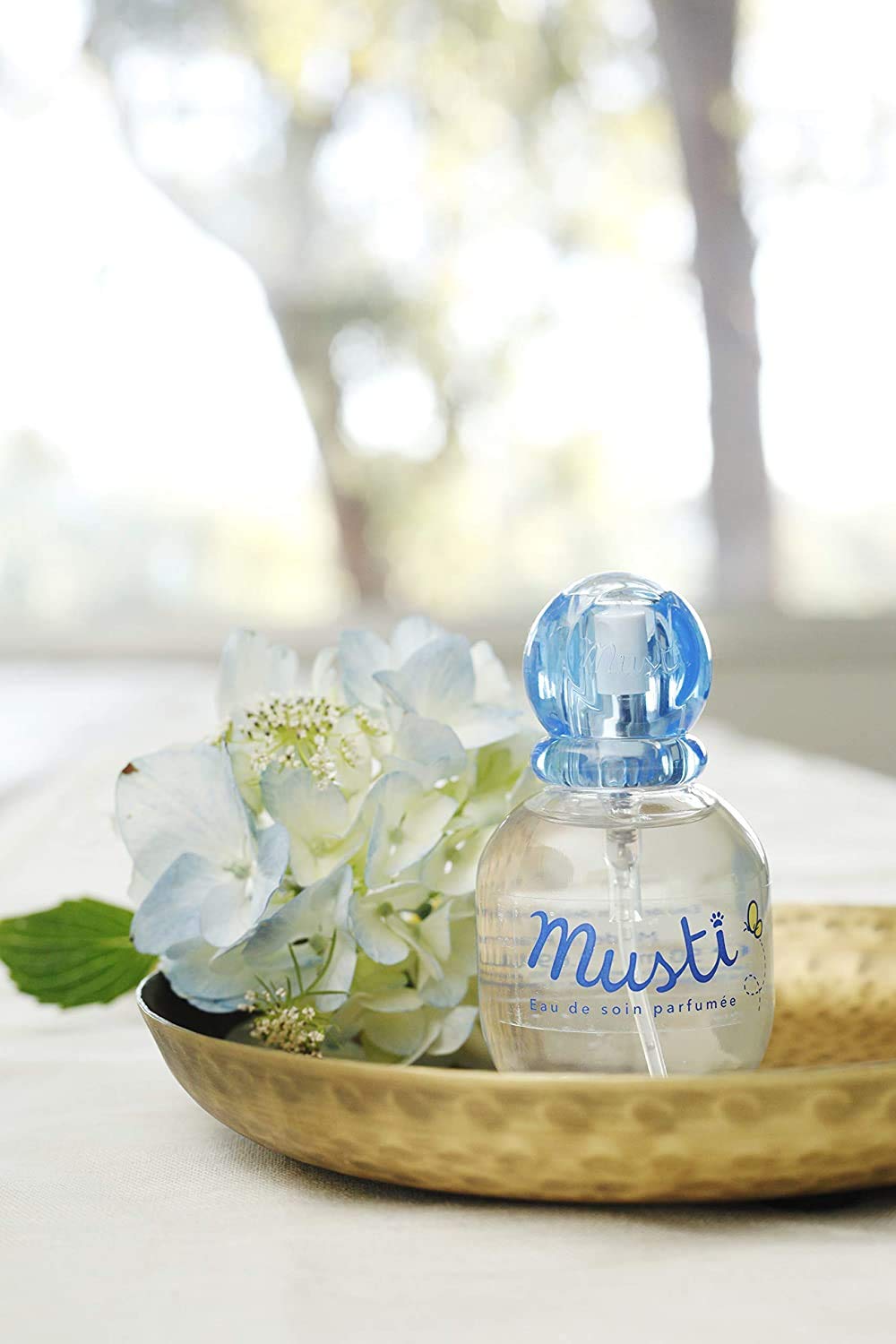Mustela perfume for babies in a clear bottle with blue cap displayed with white flowers on a brass dish