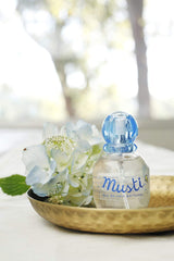 Mustela perfume for babies in a clear bottle with blue cap displayed with white flowers on a brass dish