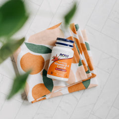 NOW bottle of Now Vitamin D3 5000 IU softgels. on orange fruit patterned cloth with marble tile background