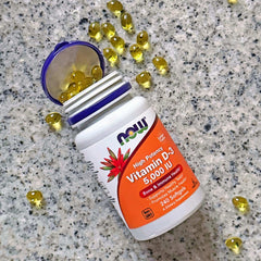 NOW vitamin D3 capsules 5000 IU softgel bottle with open purple lid and scattered golden softgels on countertop
