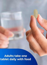 Hand holding a small yellow multivitamin tablet beside a glass of water