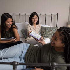Three friends on bed passing a box of Organyc organic cotton panty liners and laughing